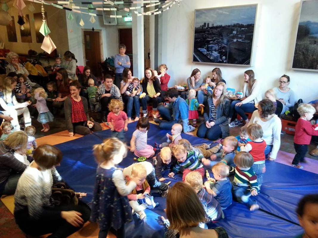 A group of young children sitting around on blue mats singing with a man with a  guitar