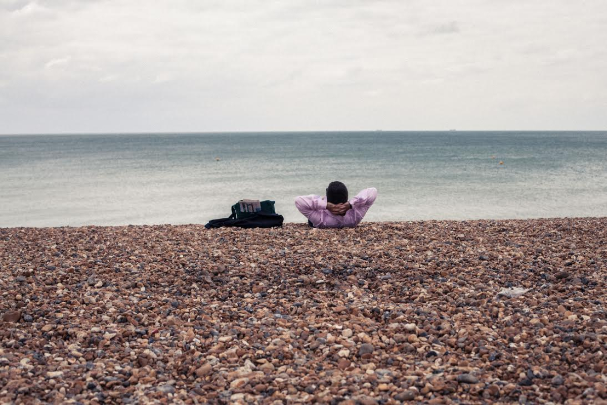 Man in pink suit and briefcase lying on a pebble beach with a blue sea horizon.
