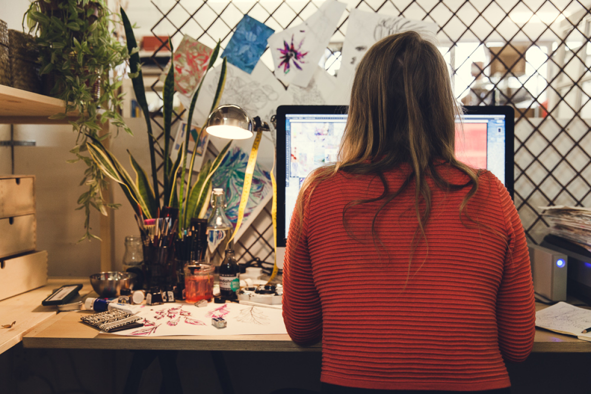 The back of a brown haired lady's head wearing an orange jumper and working at her desk with a computer screen, pen pot, plant, lamp and paintbrushes.