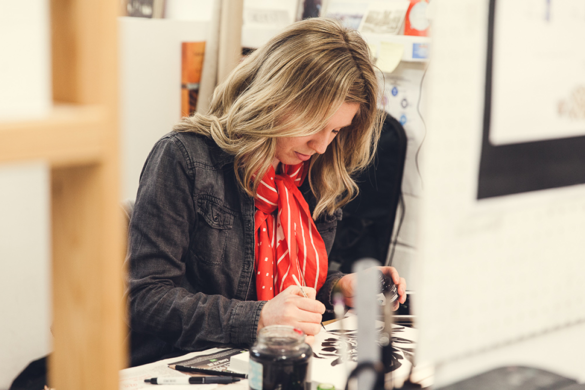 A blonde lady with a red scarf working with ink at her desk.
