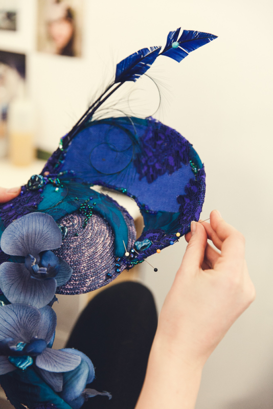 A girl in a red dress sits on a chair draped with white fur in front of a mirror whilst sewing a headpiece.