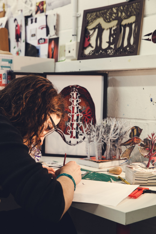 A lady with dark hair and glasses works at her desk with paper sculptures and art depicting a coral reef and a forest.