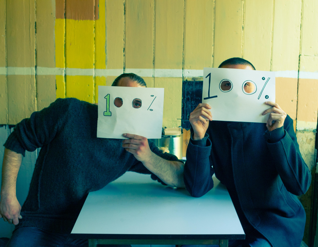 Two men wearing dark clothes, holding up pieces of paper saying one hundred percent with eye holes against a yellow and beige wooden paneled background in a cafe.