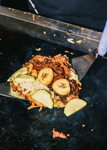 Papelon arepa being prepared on a hot plate at Netil Market, topped with black beans, fried plantain, shredded cheese, avocado slices and pulled meat