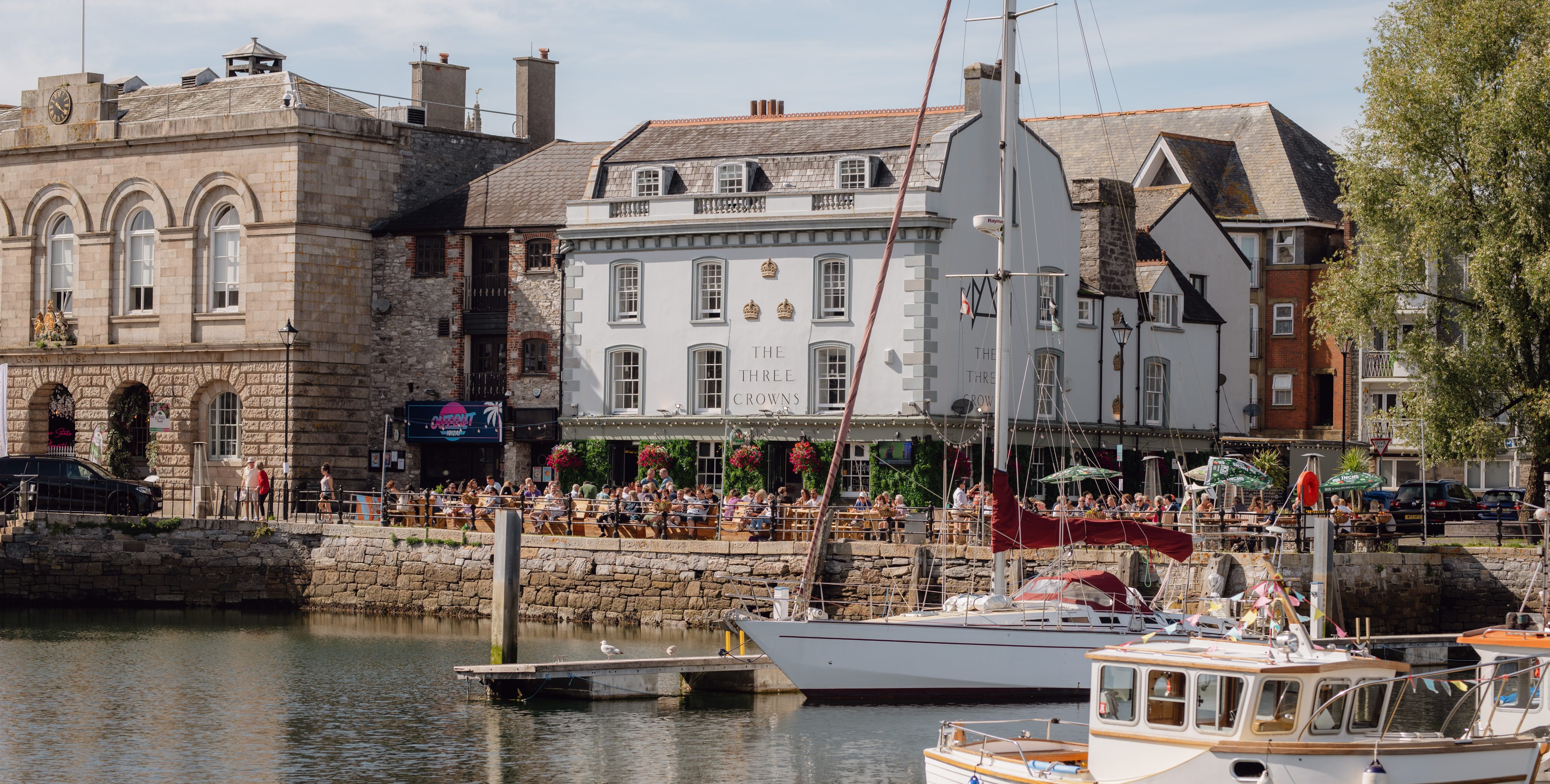 View of Plymouth’s Barbican Harbour on a sunny day, with boats docked and people dining outside, capturing the coastal charm of Eat Work Art’s Plymouth site