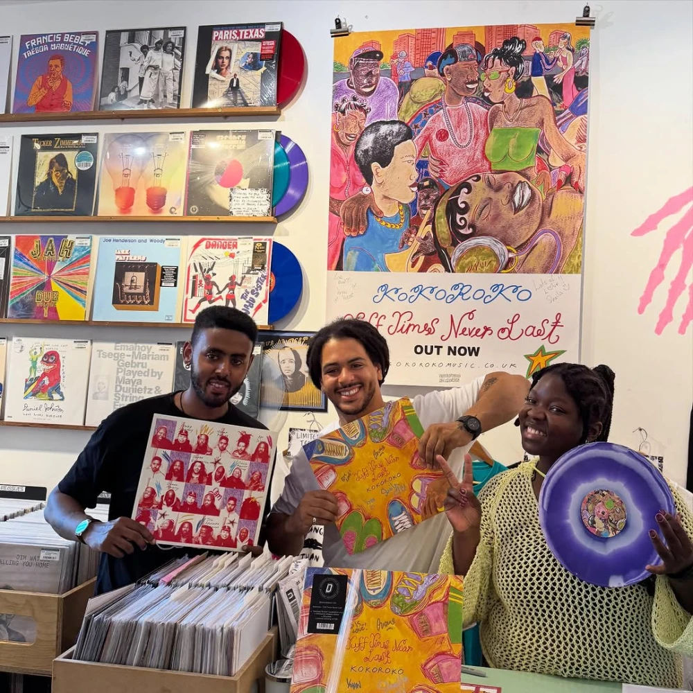 Three people holding vinyl records at Stranger Than Paradise Records, with colourful album covers and posters in the background.