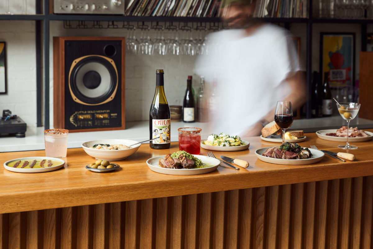 Assorted small plates, cocktails, and wine on a wooden bar counter with a blurred bartender in the background.