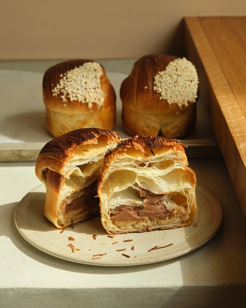 Flaky chocolate-filled pastries on a ceramic plate, with two sesame-topped pastries in the background.